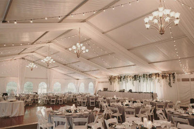 A white tent with Italian String/Market lighting, brightening up the outdoor space with white tables and chairs.