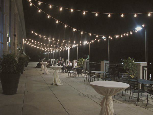 An outdoor space with tables and chairs at night adorned with Italian String/Market lighting. The lovely illumination from the lighting creates a warm and inviting ambiance.