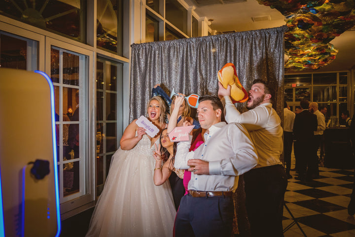 A bride and groom strike a funny pose with friends in a magic mirror style photo booth with a deluxe backdrop.