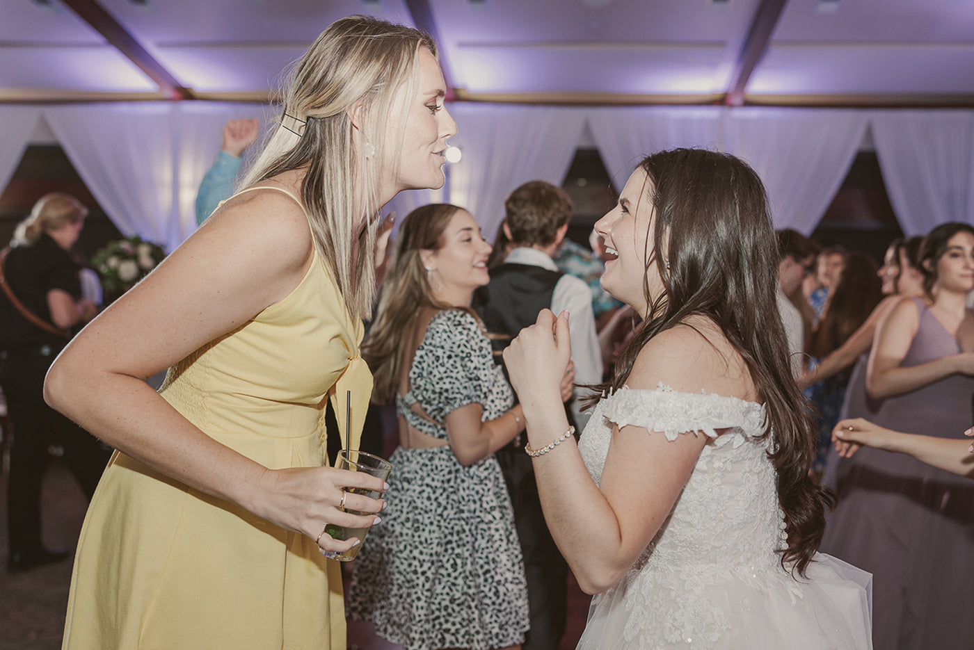 Bride and her friend grinning and dancing together amongst a full dance floor lit in purple