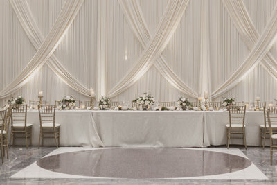 An elegant wedding reception set up with white drapes and gold chairs, featuring an exquisite straight and crossed white fabric backdrop for beautiful reception photos.