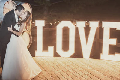 A wedding couple shares a romantic kiss in front of a huge lit LOVE marquee sign.